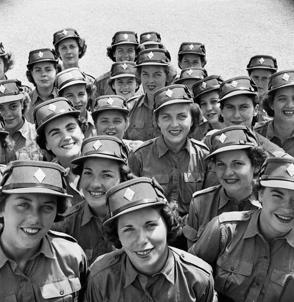 Photographie en noir et blanc – Un grand nombre de femmes arborant une casquette kaki du Service féminin de l’Armée canadienne sourient à la caméra. On voit bien les traits de leur visage.